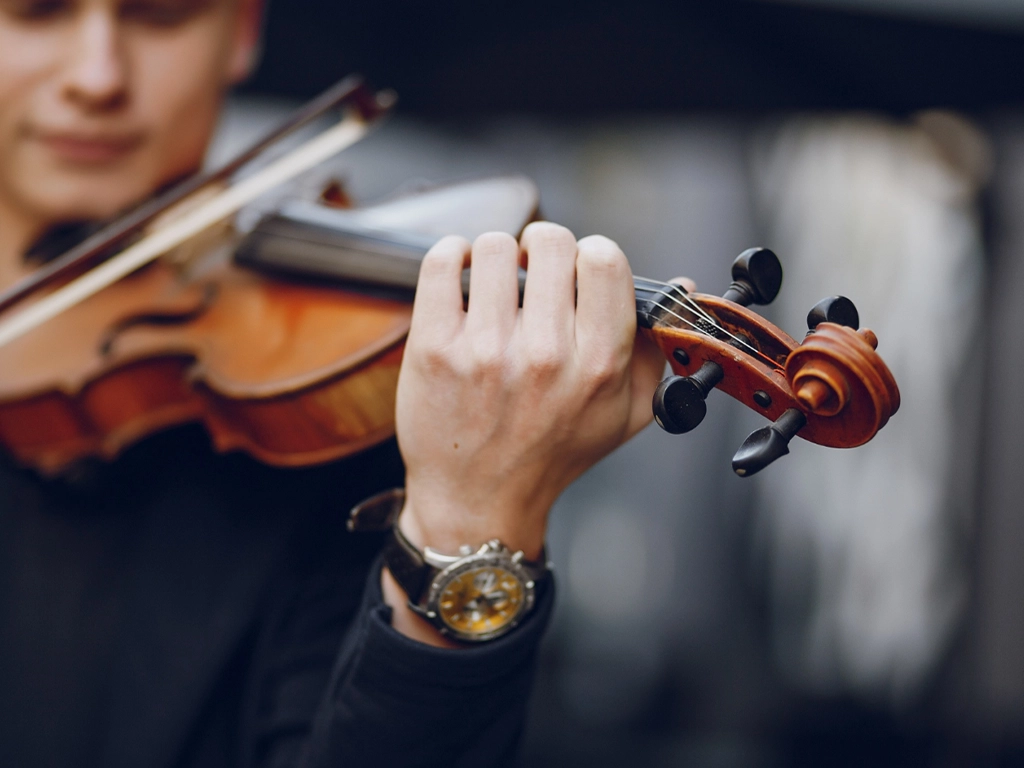 "A close-up shot of a person's hands playing a violin. The person, partially visible and blurred in the background, is wearing a black long-sleeved shirt and a large, gold-faced wristwatch. One hand holds the violin's neck and strings, while the other holds the bow, which is resting on the strings. The violin has a rich, warm brown color with visible wood grain."