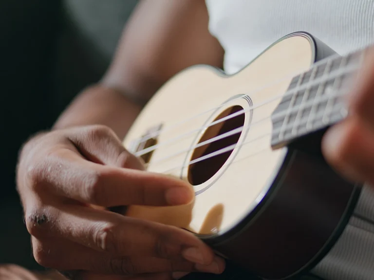 A close-up shot of a person with dark skin playing a light-colored ukulele. Their left hand is positioned on the fretboard, while their right hand is near the soundhole, strumming the strings. The person is wearing a white ribbed tank top, and the background is blurred and dark.