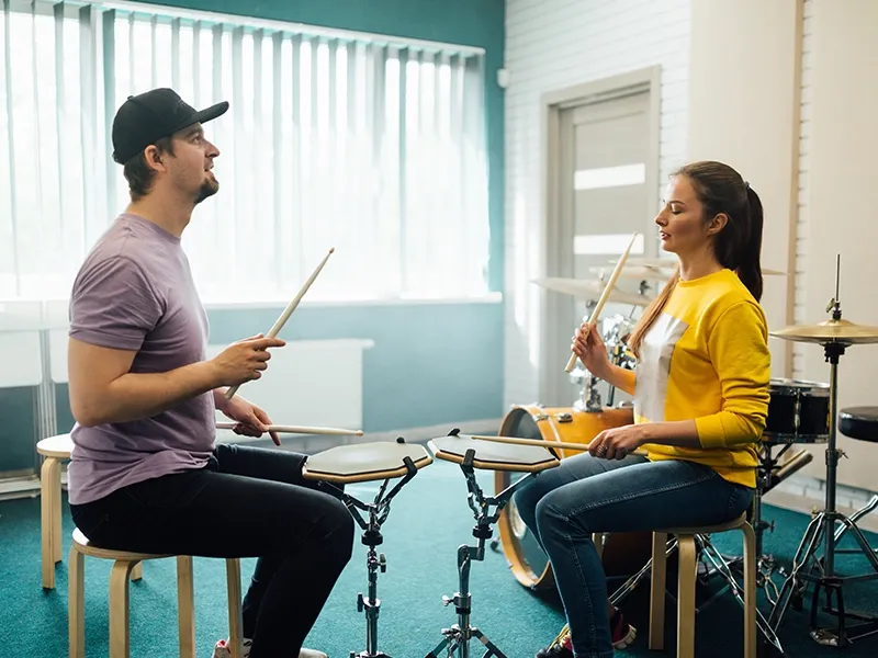 A man and a woman are seated on stools, facing each other, both holding drumsticks and playing practice drum pads. The man on the left wears a purple t-shirt and a black baseball cap, looking towards the woman. The woman on the right wears a yellow and white color-block sweater and blue jeans, looking towards the man. In the background, a full drum kit and large windows with blinds are visible, suggesting a music studio or practice room.