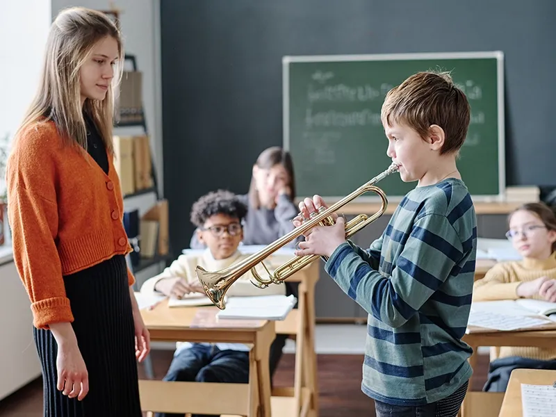 In a classroom, a young boy in a striped shirt stands, looking to his right as he plays a brass trumpet. A female teacher with long brown hair and an orange cardigan stands to his left, observing him with a focused expression. In the background, several other students are seated at desks, some looking towards the front, with a green chalkboard visible behind them.