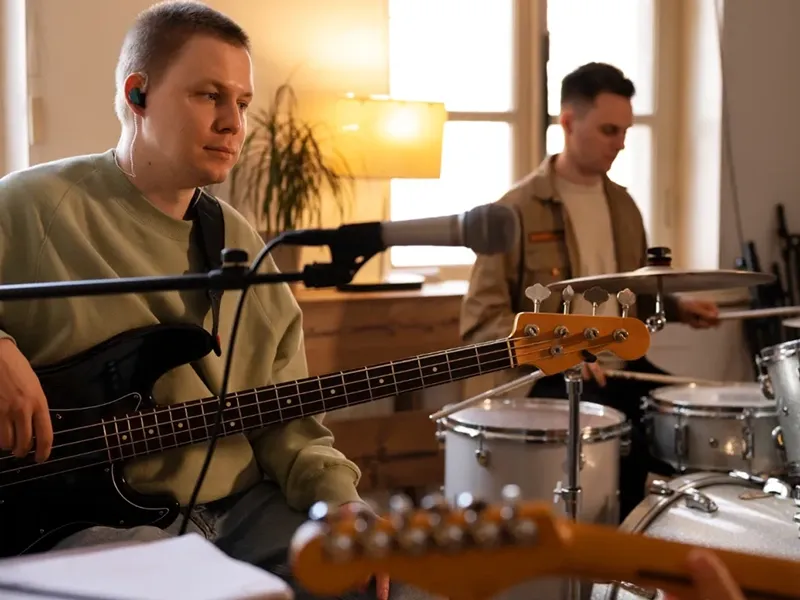 A light-skinned man in a beige long-sleeved shirt plays a black bass guitar while wearing an earbud, focused on his instrument. In the blurred background, another man plays drums, partially obscured by a microphone stand and another guitar neck in the foreground. The room has warm lighting from a lamp and sunlight.