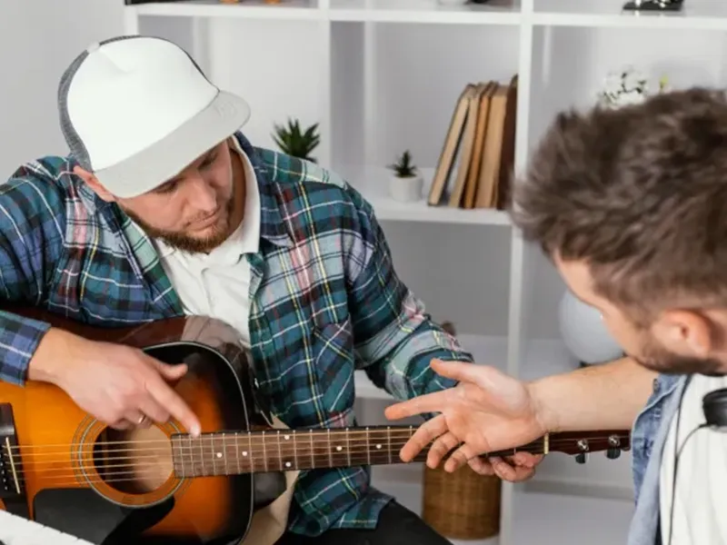 A man in a plaid shirt and baseball cap plays an acoustic guitar, looking down at the strings, while another man, partially visible, extends his hand towards the guitar's neck, observing or receiving instruction.