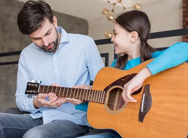 A man with a beard, wearing a light blue button-down shirt, sits next to a young girl who is playing an acoustic guitar. The man is looking down at the guitar's fretboard, seemingly instructing the girl, who is smiling and looking up at him while strumming the guitar.