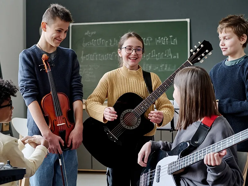Four teenage students are gathered in a music classroom with a chalkboard in the background. A girl in the center with glasses and a yellow sweater smiles while holding a black acoustic guitar. To her left, a boy in a dark sweater holds a violin. To her right, a girl with long hair and a grey sweatshirt plays a white bass guitar. Another student is partially visible on the far left. They all appear engaged and happy during their music lesson.