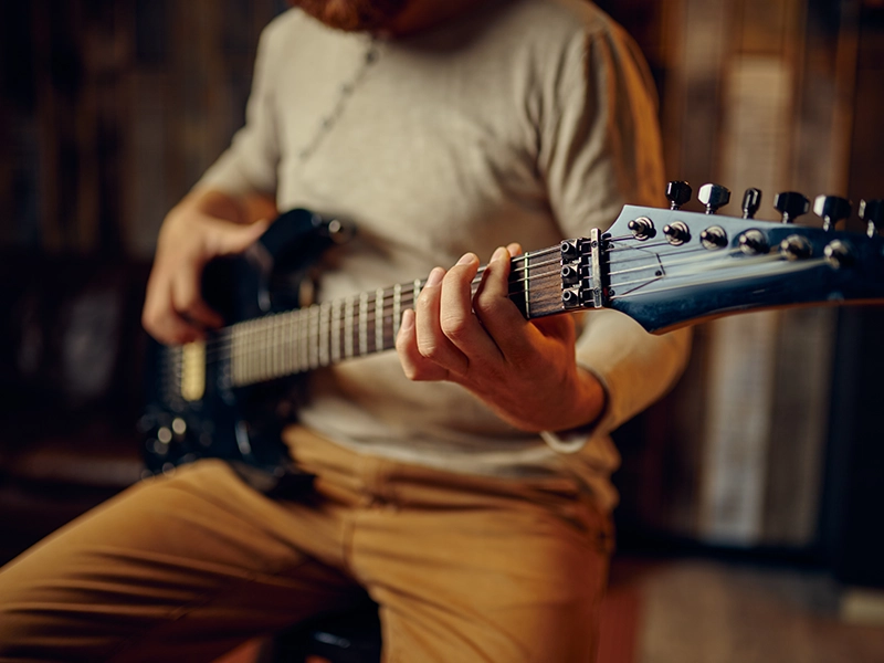 A close-up shot of a person playing a black electric guitar. The person, whose face is not visible, is wearing a light-colored long-sleeved shirt and tan pants. Their left hand is on the fretboard, pressing down on the strings, while their right hand is blurred, appearing to strum or pick near the guitar's body.