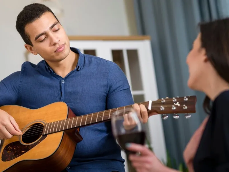 A young man with short dark hair, wearing a blue button-down shirt, is playing an acoustic guitar. His eyes are closed and his head is slightly tilted, suggesting he's engrossed in the music. To his right, partially out of frame and blurred, is a woman holding a glass of red wine, looking towards him with a smile, as if enjoying his performance. The background is a softly blurred indoor setting with a white cabinet visible.