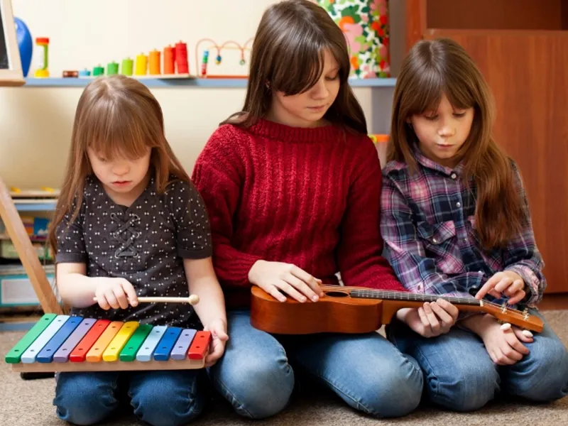 Three young girls are engrossed in playing musical instruments while sitting on the floor. The girl on the left is using a mallet to play a colorful xylophone. In the center, a girl holds a small, light-brown ukulele, her fingers poised on the strings. The girl on the right is also focused on a ukulele, adjusting something on its headstock. They appear to be in a casual, possibly home or classroom, setting, with shelves of colorful items visible in the blurred background.
