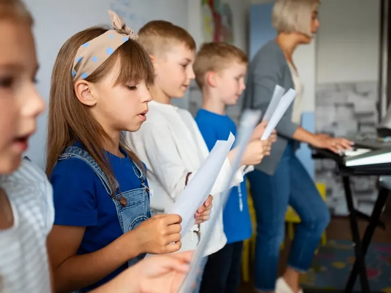 A group of diverse elementary school-aged children, led by a female teacher, are engaged in a music class. The children, spanning various ethnicities, are attentively singing while holding sheets of music, suggesting a choir or vocal practice. The teacher, positioned behind a keyboard, is either playing along or guiding them through the song. The classroom setting includes what appears to be a whiteboard or bulletin board, indicating a dedicated learning environment.