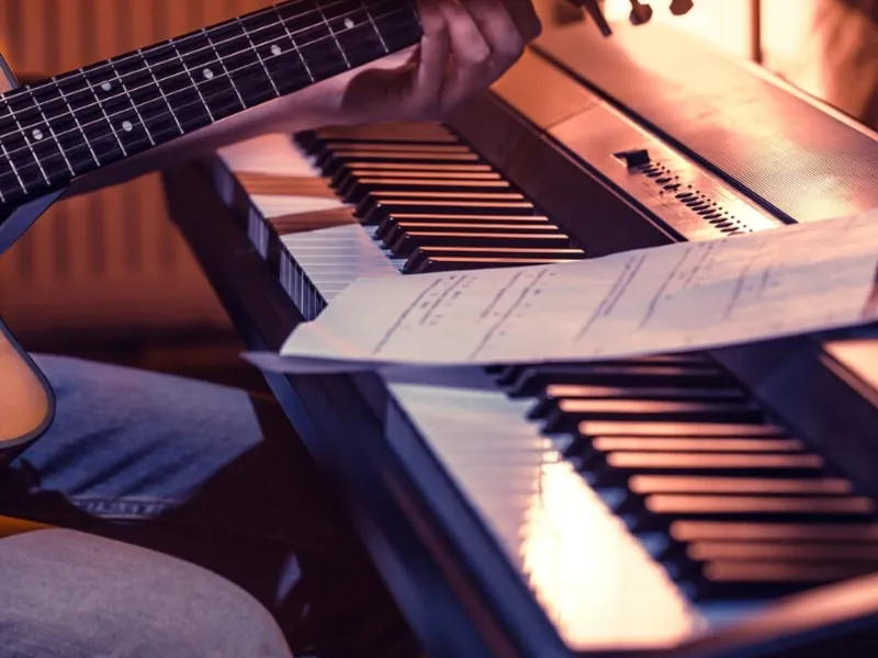 A close-up shot of a person's hands playing an acoustic guitar, with the fretboard and strings visible in the foreground. In the background, two electronic keyboards are stacked, and a sheet of music rests on the lower keyboard. The lighting suggests an indoor setting, possibly a music studio or practice space, with warm tones illuminating the instruments. The person is wearing blue jeans, partially visible at the bottom left.
