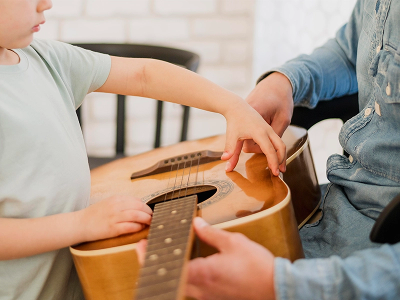 A close-up shot of a young child in a light-colored t-shirt learning to play an acoustic guitar. An adult's hands (wearing a blue denim shirt) are visible, guiding the child's fingers on the guitar's fretboard and body. The child's left hand is on the fretboard, and their right hand is touching the guitar's top near the soundhole.