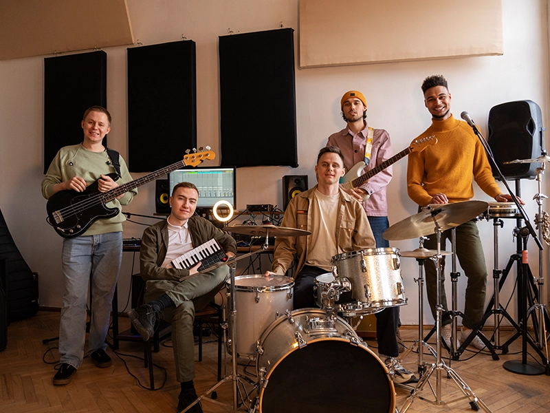 Five young adults are gathered in a music studio, posing with their instruments. From left to right: A man holds a bass guitar and smiles. Another man sits in front of him, holding a small keyboard. In the center, a man sits at a drum kit and smiles. Behind the drums, another man stands with a white electric guitar, wearing a beanie. On the far right, a man stands next to a microphone and plays a guitar. The studio features sound-dampening panels on the wall and various music equipment.