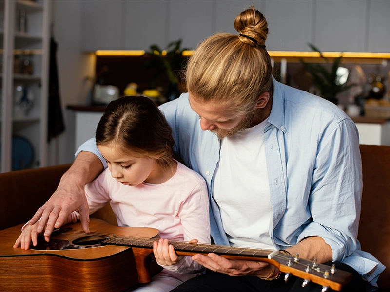 A man with long blonde hair tied in a bun and a beard, wearing a light blue shirt, sits closely beside a young girl, teaching her to play an acoustic guitar. He has his hand gently placed over hers on the guitar's strings, guiding her fingers. The girl, in a light pink shirt, looks down intently at the guitar. They are sitting on a brown couch in a warmly lit indoor setting."