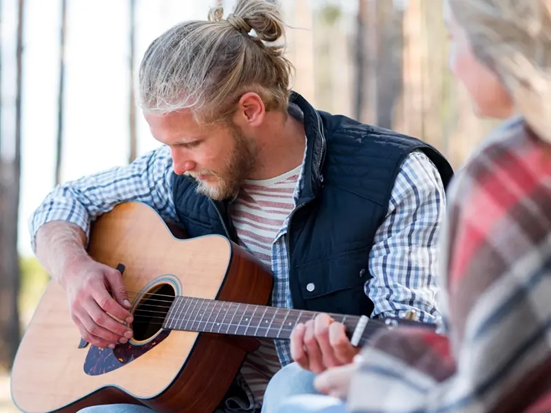 A man with blonde hair tied in a top knot and a beard, wearing a plaid shirt and a dark vest, plays an acoustic guitar. He is looking down intently at the strings. Partially visible to his right, another person wrapped in a plaid blanket is also present, likely listening. The setting appears to be outdoors, possibly in a wooded area with trees blurred in the background.
