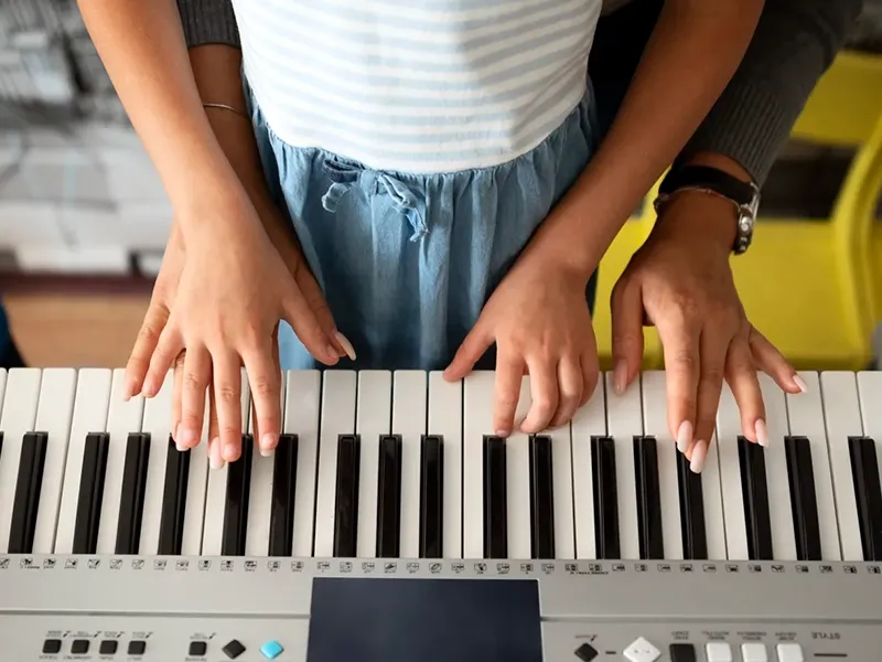 A close-up, overhead view of four hands on the keys of a digital piano. Two smaller hands, presumably a child's, are in the foreground playing the keys, while two larger hands, likely an adult's, are positioned behind and slightly over the child's hands, guiding them. The child is wearing a white and light blue striped top and a blue skirt. The adult is wearing a grey long-sleeved shirt and a watch. The background is blurred, showing light and dark elements.