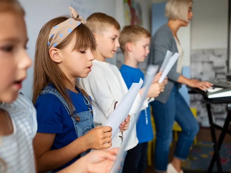 A group of young children are in a music class, standing in a line and holding sheets of music. From left to right, partially visible: a girl in a white shirt. Next to her, a girl with a light blue headband and denim overalls sings while looking at her music sheet. Behind her, two boys in white and blue shirts also hold music. In the blurred background, an adult woman with short blonde hair stands by a keyboard, seemingly leading the class.