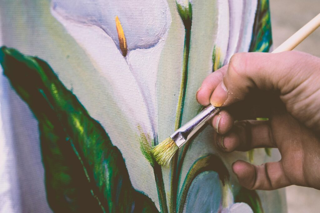 Someone paints a set of calla lilies using a thin, straight brush. The main flower is shown being painted with a large leaf to the left and more flowers on the right.