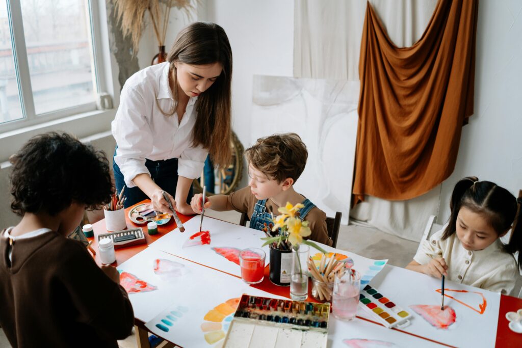 A child is sitting at a table surrounded by art supplies and paintings. He is attempting to paint a new project with the help of the teacher. The teacher is standing next to him and directing him how to move the paintbrush over the canvas.